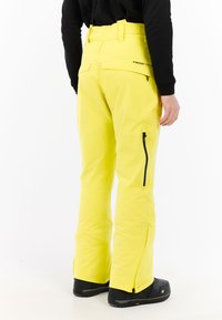 Geel waterdichte skibroek met een hoge taille, riemlussen, ritszakken aan de zijkanten en uitlopende benen, gecombineerd met zwarte schoenen.