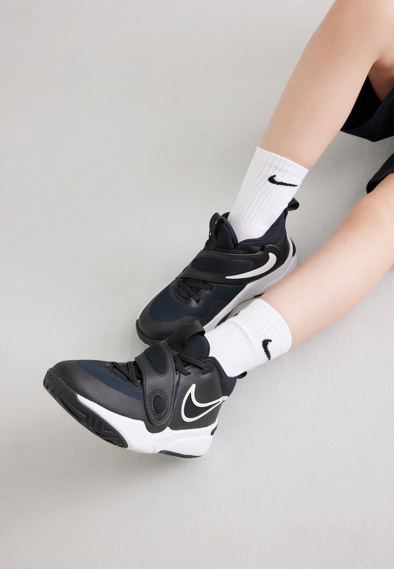Svarta Nike-sandaler med vita detaljer på ett par ben som bär vita Nike-strumpor mot en ljus, texturerad bakgrund.