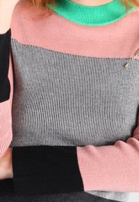 Personne portant un pull côtelé multicolore avec des manches roses, un torse gris, un col vert et des poignets noirs, les bras croisés sur la poitrine.