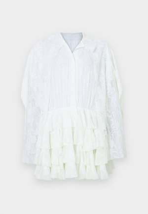 Bluza albă din dantelă cu mâneci lungi, model floral, straturi volanate în partea de jos și un guler mic, pe un fundal simplu.