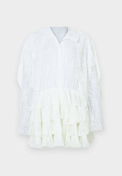 Biela čipkovaná blúzka s dlhými rukávmi, kvetinovým vzorom, vrstvenými volánmi na spodku a malým golierom, na jednofarebnom pozadí.