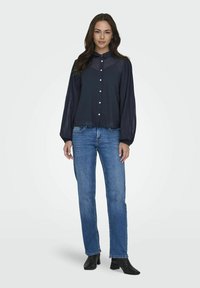 Blusa blu navy con colletto e maniche lunghe plissettate, abbinata a jeans blu a gamba dritta e stivali neri con tacco.