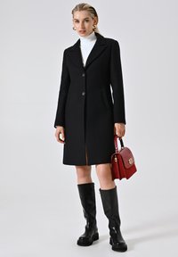 Cappotto nero su misura con rever a punta, chiusura con un bottone e due tasche laterali. Abbinato a stivali neri fino al ginocchio e una borsa rossa.