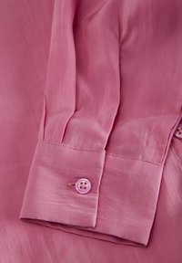 Camicia rosa a maniche lunghe con tessuto liscio, singolo bottone al polso, leggera brillantezza e dettagli cuciti con cura lungo la manica.