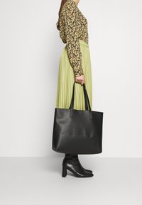 Sac fourre-tout en cuir noir à surface texturée avec double poignée, présenté aux côtés d'un chemisier à motif floral et d'une jupe plissée verte.