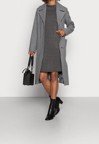 Manteau en laine grise avec revers crantés, deux poches avant et un bouton doré. Le mannequin porte une robe à carreaux grise et des bottines noires, tenant un sac noir.