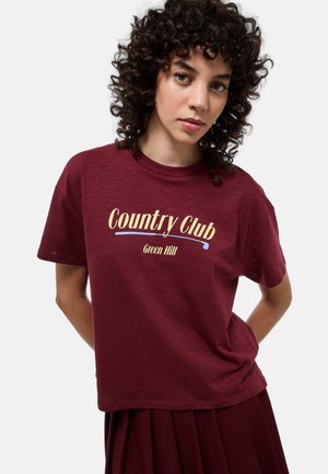 Femme aux cheveux bouclés portant un t-shirt bordeaux "Country Club Green Hill" et une jupe assortie, debout devant un fond clair uni.