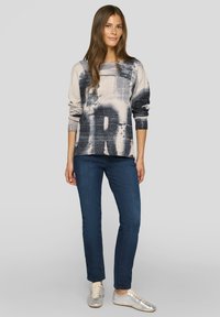 Pullover in hellem Beige mit dunkelblauem abstraktem Muster, rundem Ausschnitt, langen Ärmeln. Kombiniert mit dunkelblauen Jeans und silbernen Turnschuhen.