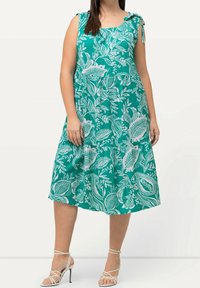 Kvinna klädd i en ärmlös teal midiklänning med vita bladmönster och axelband, kombinerat med vita högklackade sandaler med remmar.