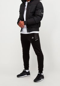 Černá péřová bunda s kapucí podšitou kožešinkou, v kombinaci s černými joggery s logem a kapsovými detaily, doplněná černými teniskami.