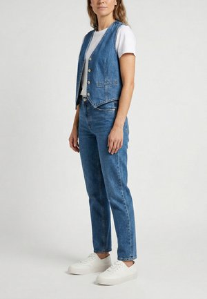 Kvinde iført hvid t-shirt, blå denim vest med knapper, matchende blå jeans og hvide sneakers, stående mod en ensfarvet baggrund.