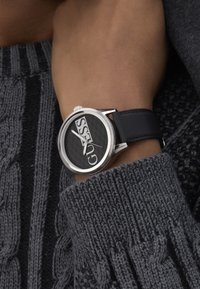 Must kollane kell, millel on suur ümar nägu, julged valged logod, hõbedased detailid ja must nahkrihm, kantuna tekstureeritud halli sviitseri peal.