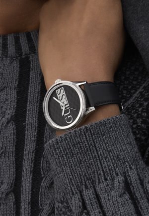 Czarny zegarek z dużą okrągłą tarczą, z wyraźnym białym logo, srebrnymi elementami oraz czarnym skórzanym paskiem, noszony na teksturowanym szarym swetrze.