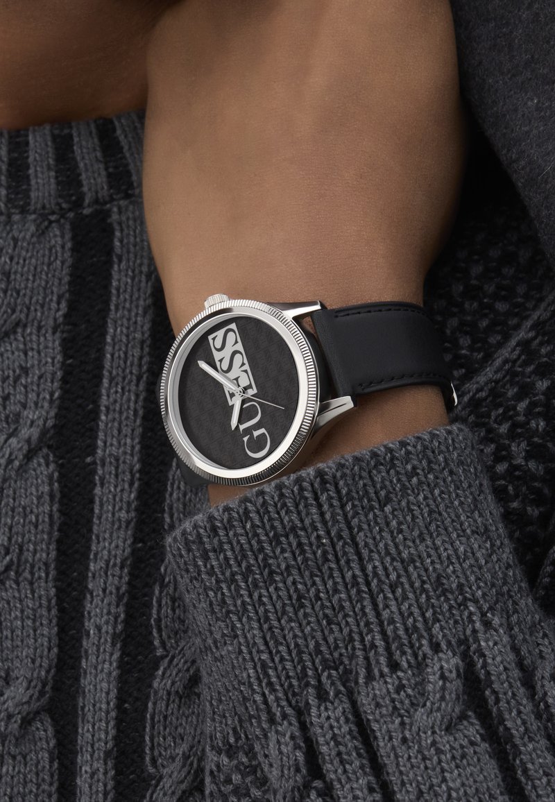 Must kollane kell, millel on suur ümar nägu, julged valged logod, hõbedased detailid ja must nahkrihm, kantuna tekstureeritud halli sviitseri peal.