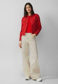 Rote Jeansjacke über einem roten Oberteil, kombiniert mit weiten, cremefarbenen Hosen. Weiche Texturen mit Knopf-Details und lässiger Passform.