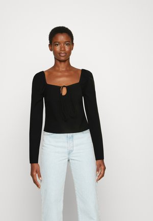 Top preto de mangas longas com decote quadrado e detalhe de laço ao centro. Apresenta mangas ajustadas e uma textura suave. Combinado com jeans azul claro.