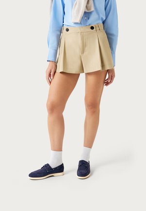 Persoon draagt beige geplooide shorts met zwarte knopen, een lichtblauw overhemd, witte sokken en marineblauwe bootschoenen, staand tegen een effen achtergrond.