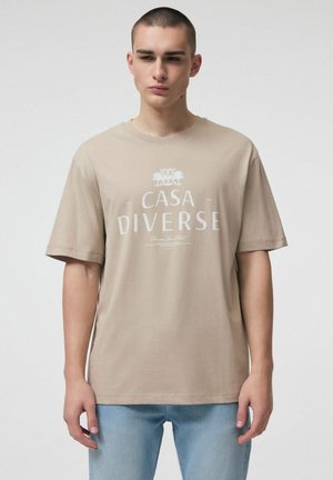 Młody mężczyzna w beżowej koszulce "CASA DIVERSE" i jasnoniebieskich dżinsach, stojący na tle jednolitego, jasnego tła.