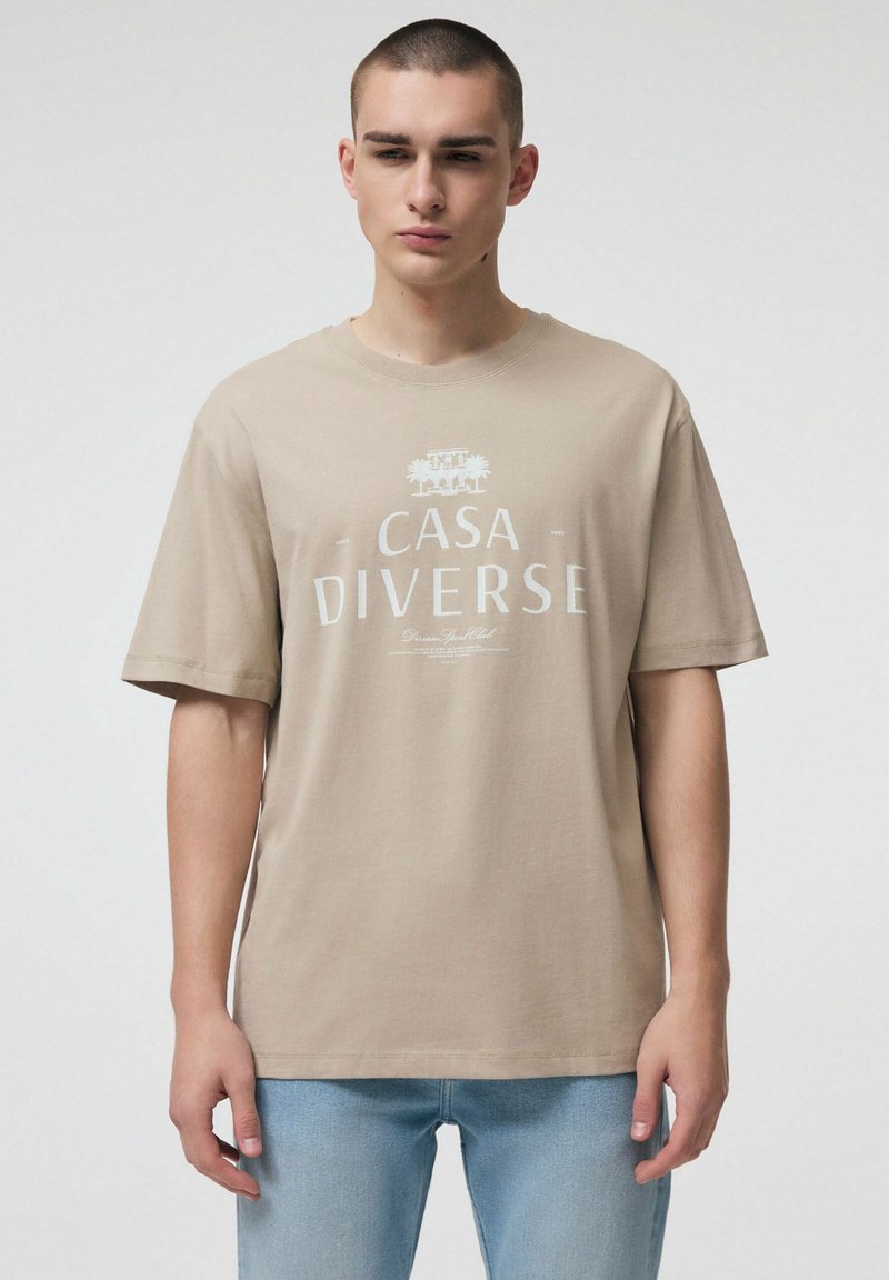 Młody mężczyzna w beżowej koszulce "CASA DIVERSE" i jasnoniebieskich dżinsach, stojący na tle jednolitego, jasnego tła.