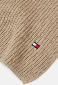 Beige strikket hat med ribbet tekstur; inkluderer et lille broderet logo med rød, hvid og blå farver på den ene side.