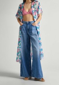 Kvinna klädd i blommig kimono, rosa bralette, högmidjade vida jeans och vävda sandaler, stående med händerna i fickorna.