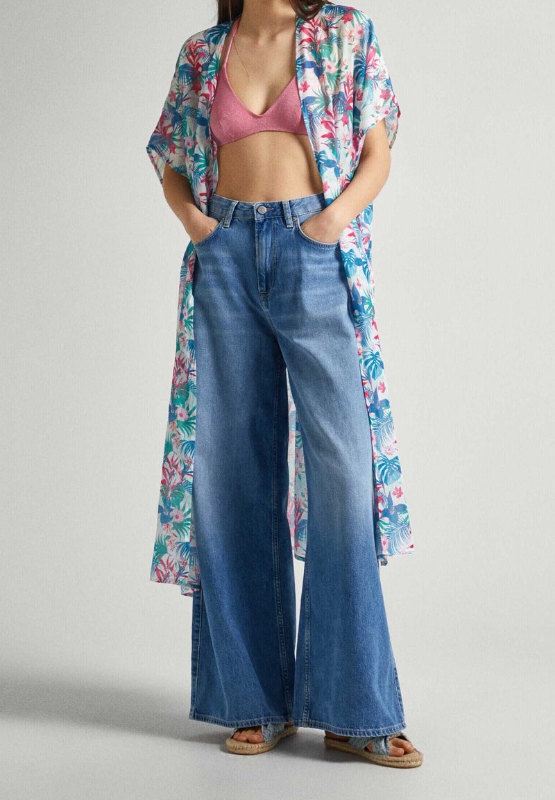 Kvinna klädd i blommig kimono, rosa bralette, högmidjade vida jeans och vävda sandaler, stående med händerna i fickorna.