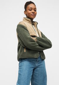 Μπλούζα φλις σε σκούρο πράσινο με επάνω τμήμα από μπεζ νάιλον, με ψηλό γιακά, κλείσιμο με φερμουάρ και πλαϊνές τσέπες. Λεπτομέρεια υφής υφάσματος.