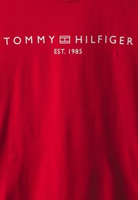 Rotes Baumwoll-T-Shirt mit weißem Text, der "TOMMY HILFIGER EST. 1985" zeigt, mit einem Box-Logo, glatter Textur und Rundhalsausschnitt.