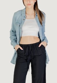 Camisa de mezclilla ligera sobre un top corto blanco de canalé, combinada con pantalones negros de cintura elástica. Con botones en el frente, mangas largas y bolsillos visibles.