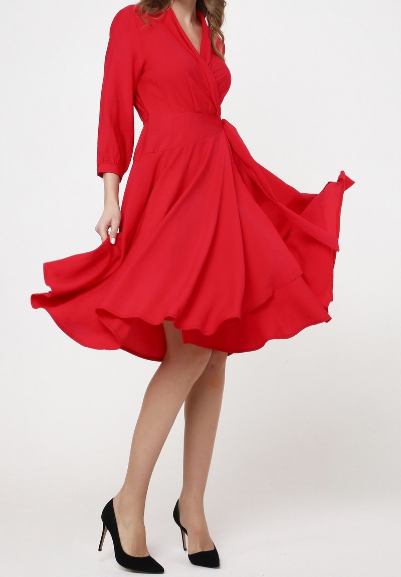 Robe portefeuille rouge avec manches trois-quarts, jupe évasée et liens à la taille. Le tissu semble léger et lisse. Chaussures à talons hauts noires.
