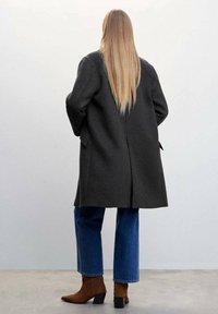 Grijze wollen jas met een rechte ontwerp, grote achteropening en zijzakken. Gecombineerd met blauwe denimjeans en bruine enkellaarsjes.