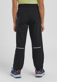 Pantalons softshell noirs avec taille élastique, bandes réfléchissantes et poche latérale zippée ; associés à des chaussures bleues et oranges.