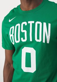 Personne portant un t-shirt vert avec un texte blanc "BOSTON" et le chiffre zéro, avec un logo Nike sur la partie supérieure de la poitrine.