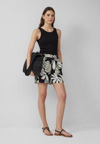 Débardeur noir à col rond, short noir et blanc à motif de feuilles tropicales, et sandales plates noires. Sac en toile noir à la main.