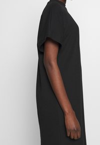 Profil d'une personne portant une robe noire à manches courtes et ample devant un fond blanc uni.