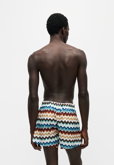 Homme à la peau foncée et aux cheveux courts et bouclés, debout de dos, portant un short coloré à motif zigzag sur un fond clair uni.