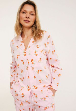 Vrouw draagt lichtroze pyjama met sinaasappelprint, staat met handen in de zakken tegen een effen achtergrond.
