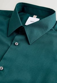 Dunkelgrünes Hemd mit Knöpfen aus strukturiertem Stoff, das einen spitzen Kragen, ein weißes Etikett und graue Knöpfe an der Knopfleiste aufweist.