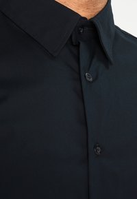 Granatowa koszula na guziki z klasycznym kołnierzykiem, gładką fakturą i czarnymi plastikowymi guzikami. Zawiera widoczne detale szycia na kołnierzyku.