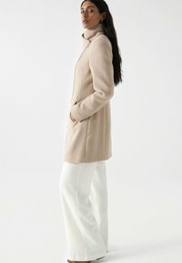Manteau en laine beige avec un col montant, fermeture éclair à l'avant et poches latérales, porté sur un pantalon blanc évasé. Texture lisse et coupe ajustée.