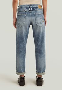 Jean en denim bleu clair avec une coupe décontractée, bords roulés, deux poches arrière et une patch en cuir brun sur la ceinture.