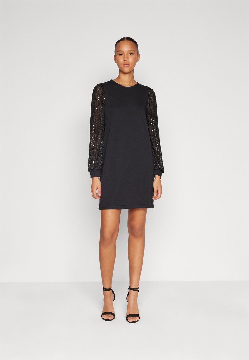 Robe sweat noire avec de longues manches en sequins. Dispose d'un col rond et d'une coupe décontractée, associée à des talons noirs à lanières.