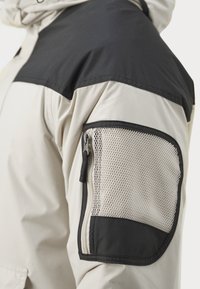 Beige jakke med sorte skulderpaneler, som har en glidelås mesh-lomme på ermet. Materialet ser glatt ut med en lett glanset finish.