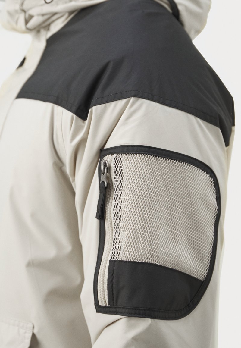 Beżowa kurtka z czarnymi panelami na ramionach, z zamykaną sieciową kieszenią na rękawie. Materiał wydaje się gładki z lekko błyszczącym wykończeniem.