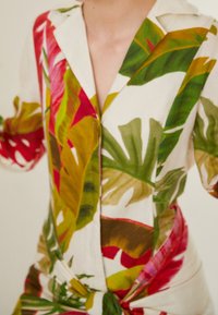 Blumenleinenhemd mit lebhaften grünen, roten und gelben Blattmustern, einem eingekerbten Kragen und einem gebundenen Taillendetail, das ein entspanntes Design zeigt.
