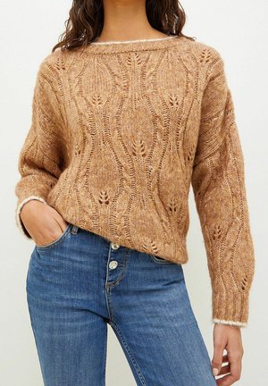 Kvinde iført en tanfarvet kabelstrikket sweater med balloneærmer, kombineret med højtaljede blå jeans med knapper, hånd i lommen.