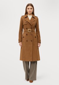 Brun dubbelknäppt trenchcoat med bältad midja, axelband och knappdetaljer, tillverkad av ett texturerat tyg.