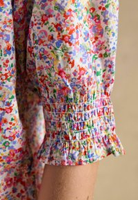 Manche en tissu fleuri avec des fleurs rouges, violettes, vertes et bleues rassemblées au niveau du poignet par un smock élastique et une petite bordure à volants.