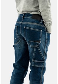 Pantalones cargo de mezclilla en azul oscuro con múltiples bolsillos, costuras visibles y una textura suave. Presenta una etiqueta de logo negra en la cintura.