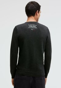 Schwarzer Strickpullover mit strukturierter Oberfläche, versehen mit einem gerippten Saum und Bündchen. Auf der Rückseite ist weißer und roter gedruckter Text zu sehen.